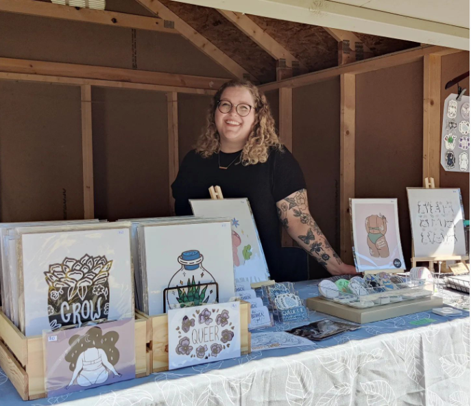 Jess standing behind a table of body positive, planty, and queer prints at a Market in Kitchener.